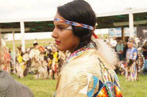 A young Native American teen wearing ceremonial dress at a Pow Wow observing the activities.