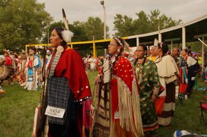 A line of Native American adults dressed in full ceremonial dress at a Pow Wow.